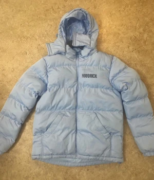 Ljusblå pufferjacka från Hoodrich - Säljer en ljusblå pufferjacka från Hoodrich med stor logga på ryggen och mindre logga på bröstet. Jackan har huva, två stora fickor framtill och dragkedja. Perfekt för kalla dagar och har en riktigt chill vibe.