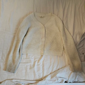 Ljusbeige stickad kofta med knappar - Ljusbeige stickad kofta från Kappahl med v-ringad hals och tre stora knappar framtill. Koftan är croppad och har långa ärmar. Storlek 146-152