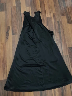 Svart långklänning med omlott - Snygg svart långklänning med omlott-detalj framtill och ärmlös design. Klänningen har en stilren och enkel look med lätt glansigt tyg som ger en elegant känsla. Perfekt för dig som gillar minimalistisk stil och vill ha något som sticker ut utan att vara för mycket.