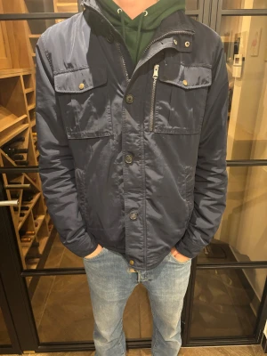 J.lindeberg jacka mörkblå  - Snygg mörkblå vinterjacka från j.lindeberg med flera praktiska fickor framtill, både med knapp och dragkedja. Jackan har hög krage, långa ärmar och knäppning med både knappar och dragkedja. Perfekt för kallare väder. Storleken är xl men person på bilden är 188 cm och skulle säga mer åt M/L hållet. 