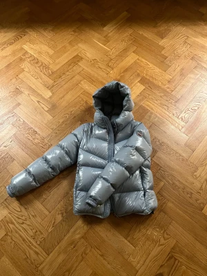 Ljusblå Colmar pufferjacka med huva - Snygg grå pufferjacka från Colmar med glansig finish och huva. Jackan har en quiltad design, dragkedja framtill och ribbade muddar vid ärmslut. Perfekt för kalla dagar och har en modern, oversized look. Colmar-logga på ärmen och insidan.