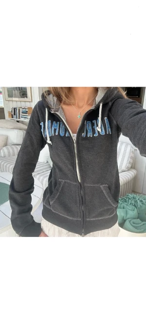 Mörkgrå Abercrombie hoodie med dragkedja - Snygg mörkgrå hoodie från Abercrombie med blått broderat tryck över bröstet. Tröjan har huva med snörning, två fickor fram och dragkedja.