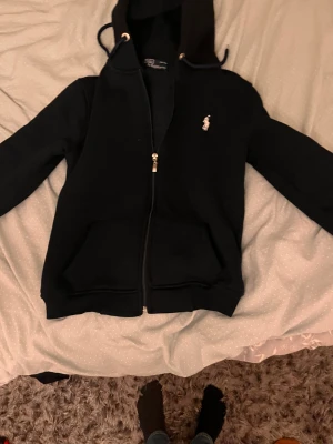 Svart hoodie från Polo Ralph Lauren - Svart hoodie med dragkedja från Polo Ralph Lauren. Klassisk design med vit broderad logga på bröstet, två fickor fram och justerbar huva med snören. Mjuk insida och ribbade muddar. Perfekt för en chill och stilren look. Står 158 men är ungefär 146-152