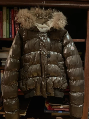 Brun Moncler Pufferjacka med Päls - Vintage brun pufferjacka med pälsluva från Moncler. Jackan har både knappar och dragkedja framtill. Bra till vintern då den har skön och tjock fyllning och värmer bra. Den har en väldigt lätt och nästan osynlig missfärgning på ena armen men har dock fläckar på insidan, övrigt skick är bra.                Storlek: 1 (S) Jackan är perfekt som julklapp till flickvän eller syskon.