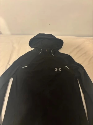Svart vindjacka från Under Armour - Svart vindjacka från Under Armour med huva och hel dragkedja framtill. Jackan har vita detaljer vid axlarna och logga på bröstet samt stor Under Armour-text på ryggen. Perfekt för träning och aktiva dagar, tillverkad i lätt polyester. Knappt använd då den är lite för stor på mig