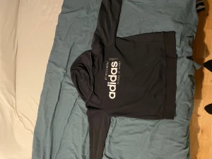 Svart hoodie från Adidas Junior XL - Svart hoodie från Adidas i Junior storlek XL med stor vit logga och texten 'Code of Sport' på bröstet. Klassisk modell med huva och dragsko, ribbade muddar och mjukt material i bomullsblandning. Perfekt för en sportig och avslappnad stil.