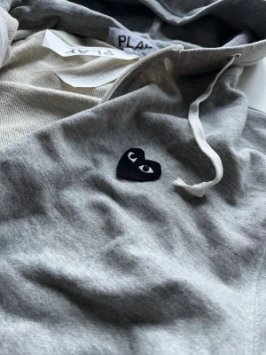 Grå hoodie Comme des Garçons Play - Grå zip-hoodie från Comme des Garçons Play med svart hjärta med ögon broderat på bröstet. Klassisk huva med vita snören, två fickor fram och ribbade muddar. Tillverkad i 100% bomull för en skön och avslappnad vibe.