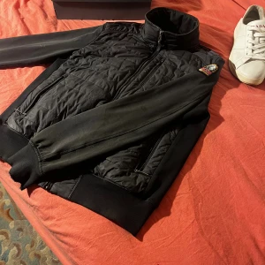 Svart pufferjacka från Parajumpers - Svart pufferjacka från Parajumpers med quiltad front och rygg, släta svarta ärmar och ribbade muddar. Jackan har hög krage, dragkedja och flera fickor med zip. Insidan är ljus och har märkets patch och orange detalj. Perfekt för kalla dagar.