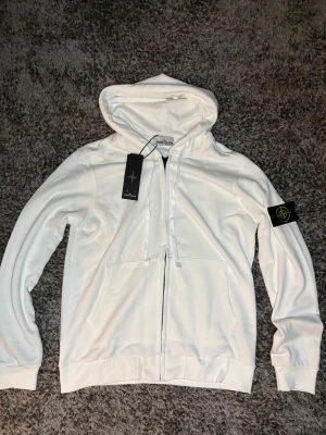  Stone Island - Vit hoodie från Stone Island med klassisk logopatch på vänster ärm. Tröjan har huva med snörning, dragkedja framtill och två fickor. Materialet är mjuk bomull och passformen är normal. Perfekt för dig som gillar streetwear och snygga detaljer.