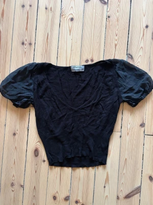 Svart blus från NOANOA med puffärm - Svart blus från noanoa med v-ringning och puffiga kortärmar i tunnare, prickigt tyg. Blusen är mjuk och stretchig, medan ärmarna har en lätt transparent känsla. Perfekt för dig som gillar en feminin och cool stil.