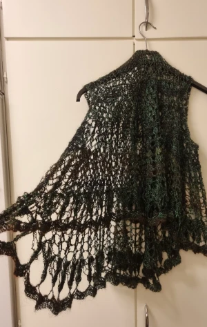 Handstickad mörkgrön poncho i ull - Unik handstickad poncho mörkgrönt och svart med vackert spetsmönster. Sjalen har en luftig och böljande form med fina detaljer längs kanterna. Perfekt att svepa runt axlarna för en extra touch till din outfit.