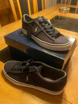 Mörkblå sneakers från Polo Ralph Lauren - Snygga mörkblå sneakers från Polo Ralph Lauren med klassisk design. Skorna har vit platt sula, rund tå och snörning. Materialet är canvas och de har Polo-logga broderad i guld på sidan samt tryckt på hälen och plösen. Perfekta för en chill och stilren look.