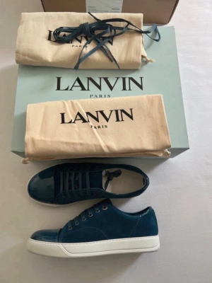 Mörkblå sneakers från Lanvin - Säljer ett par snygga mörkblå sneakers från Lanvin med vit platt sula och snörning. Skorna har en stilren design med blank tå och ovandel i mocka. Perfekta för dig som vill ha en exklusiv och modern look.