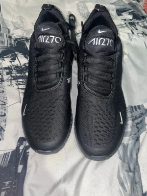 Nike Air Max 270 svarta sneakers - Svarta Nike Air Max 270 sneakers med vit logga och detaljer. Ovandel i mesh och syntet, snörning och rund tå. Sulan har vita prickar och synlig Air-enhet på hälen. Perfekta för dig som gillar sportig stil och vill ha bekväma skor med snygg design.