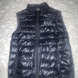 Svart dunväst från EA7 Emporio Armani - Säljer en svart, quiltad dunväst från EA7 Emporio Armani med glansig finish och logga på bröstet. Västen har ståkrage och dragkedja framtill. Perfekt för lager-på-lager och har en modern, sportig look.