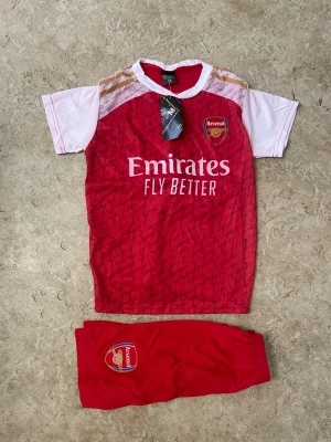 Arsenal matchställ G.Jesus nr 9 - Säljer ett Arsenal matchställ med tröja och shorts. Tröjan är röd med vita ärmar, guldiga detaljer och klubbmärke på bröstet. På ryggen står G.JESUS och nummer 9. Shortsen är röda med Arsenal-logga. Perfekt för fotbollsfans!