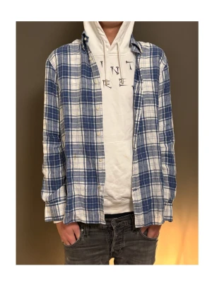 Blåvit rutig skjorta i bomull - Säljer en blå och vit rutig skjorta i mjuk bomull. Skjortan har klassisk krage, knäppning framtill och långa ärmar med manschett. Perfekt att bära öppen över en hoodie eller stängd för en mer clean look.