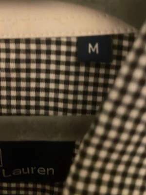 Rutig skjorta från Polo Ralph Lauren - Snygg svartvit rutig skjorta från Polo Ralph Lauren i storlek M. Skjortan har klassisk krage, knappar framtill och det ikoniska broderade Polo-logot på bröstet. Tillverkad i mjuk bomull som är skön mot huden och passar perfekt till jeans eller chinos.