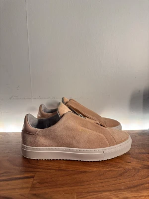 Rosa Axel Arigato Sneakers - Snygga rosa sneakers från Axel Arigato i mjuk mocka med vit platt sula och cool dragkedja framtill istället för klassisk snörning. Minimalistisk design med rund tå och diskret logga i guld på sidan. Perfekta för dig som gillar stilrena och moderna skor.