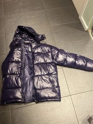 Mörkblå dunjacka från Moncler - Säljer en mörkblå dunjacka från Moncler med glansig finish och huva. Jackan har långa ärmar, dragkedja framtill och fickor med dragkedja. Klassisk Moncler-logga på ärmen. Perfekt för kalla dagar och riktigt snygg stil.