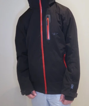 Svart vindjacka från North Bend - Svart vindjacka från North Bend med huva och snygga röda dragkedjor. Jackan har flera fickor med dragkedja, långärmad design och justerbara muddar. Perfekt för dig som gillar att vara ute och vill ha en stilren jacka med coola detaljer.
