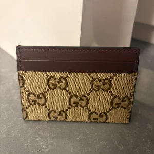 Gucci plånbok i beige och brunt - Säljer en snygg plånbok från Gucci med klassiskt GG-mönster i beige och brunt. Övre delen är i mörkbrunt skinn och resten i canvas med logotyptryck. Perfekt för kort och enkel att ha med sig i fickan.