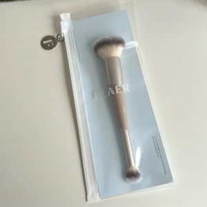 FLAER foundation- och concealerborste - Snygg duo-sminkborste från FLAER med syntetiskt hår, perfekt för applicering av foundation och concealer. Borsten har ett fluffigt, ljust borsthuvud med mörka toppar och ett stilrent handtag i beige och silver. Handtaget är gjort av återvunnen aluminium och plast. Ordinarie 229kr pris 170kr