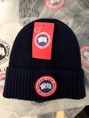 Canada Goose stickad mössa marinblå, grå, vit - Säljer en ribbstickad marinblå mössa från Canada Goose med klassisk rund patch framtill. Mössan har uppvikt kant och är riktigt mjuk och skön. Perfekt för kalla dagar och snygg till jackan.