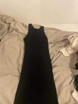 Svart långklänning från 157 - Svart ribbad långklänning från 157 i storlek XS. Klänningen är ärmlös och har en rund halsringning. Tillverkad i 100% bomull vilket gör den mjuk och skön att bära. Perfekt för dig som gillar enkel och stilren design.