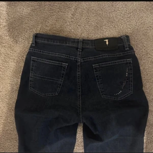 Mörkblå bootcut jeans med detaljer - Säljer ett par mörkblå jeans från Trussardi Jeans med bootcut-snitt. Jeansen har klassiska fem fickor, kontrastsömmar och snygga detaljer och broderi på bakfickan. Materialet är jeans och passformen är normal. Perfekt för dig som gillar stilrena jeans med lite edge.