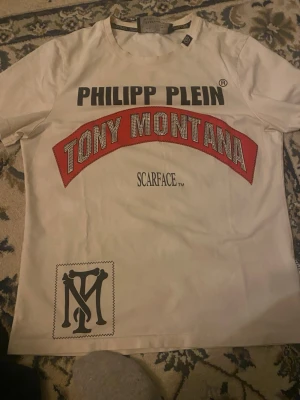 Vit Philipp Plein Scarface t-shirt - Säljer en vit t-shirt från Philipp Plein Platinum Cut med stor print på bröstet: 'Tony Montana' i rött och 'Scarface'. Coolt tryck och svart logga nertill. Perfekt för dig som gillar statement-plagg och filmreferenser.