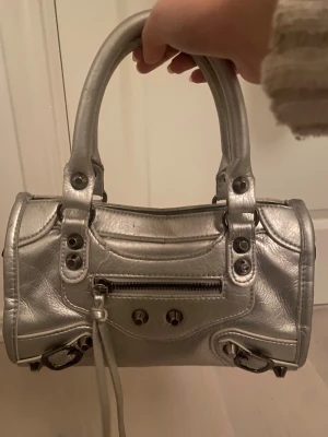 Silver handväska med dragkedja - Cool handväska i metallic silver med två rejäla handtag och snygga metalldetaljer. Väskan har en praktisk dragkedja framtill och dekorativa nitar. Tillverkad i skinnimitation som ger en lyxig känsla. Perfekt storlek för det viktigaste och riktigt trendig look.