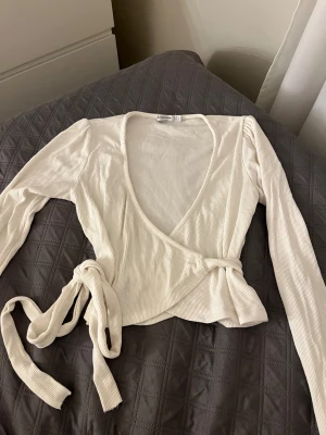 Vit omlottopp med knytband - Säljer en vit omlottopp från Stradivarius. Toppen har långa ärmar, v-ringad hals och knytband i midjan som ger en snygg siluett. 