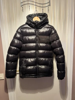 Svart pufferjacka från Alessandro Zavetti - Snygg svart pufferjacka från Alessandro Zavetti med huva och feta, quiltade paneler. Jackan har dragkedja framtill, två sidofickor med dragkedja och en diskret logga på bröstet. Materialet är glansigt syntet och jackan är perfekt för kalla dagar. Liten defekt då bokstäverna börjar lossna lite annars mycket bra skick, nypris runt 1400kr mitt pris 599kr kom gärna med pris förslag !