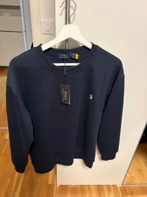 Mörkblå sweatshirt från Polo Ralph Lauren - Snygg mörkblå sweatshirt från Polo Ralph Lauren i bomull. Klassisk rund hals och långärmad modell med den ikoniska broderade loggan i vitt på bröstet. Perfekt för dig som gillar stilrena och tidlösa plagg med premiumkänsla. Storlek M, helt ny, aldrig använd! Priset kan diskuteras!