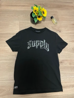 Svart Supply & Demand t-shirt - Svart t-shirt från Supply & Demand med tryck i silver på bröstet och en stor grafisk ängel med text på ryggen. Klassisk rund hals och korta ärmar. Materialet är mjuk bomull som känns skönt mot huden. Perfekt för dig som gillar streetwear och coola prints.