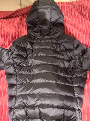 Svart pufferjacka från Colmar, strl 44 - Säljer en svart pufferjacka från Colmar med huva och snyggt quiltad design. Jackan har en silverfärgad dragkedja framtill och Colmar-logga på ärmen. Insidan är ljusgrå och materialet är polyester, perfekt för kalla dagar. Kolla alla bilder för se hur den ser ut skick 10/10 jätte bra självklart äkta 