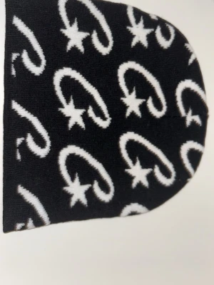 Cortez mössa - Cool svart stickad väst med vitt mönster av stjärnor och böjda linjer. Västen har en rundad form och är tillverkad i ett mjukt stickat material som känns skönt mot huden. Perfekt för dig som gillar grafiska detaljer och vill sticka ut lite extra.