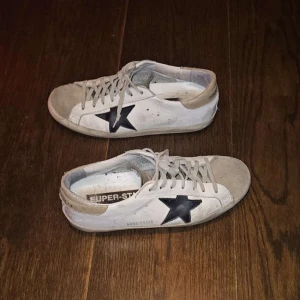 Golden Goose Superstar sneakers vit/svart - Golden Goose Superstar sneakers i vitt med svart stjärndetalj på sidan och grå mocka vid tån. Skorna har snörning, rund tå och platt sula. Beige hälparti och klassisk logga på insidan. Levereras med originalkartong och dustbag.