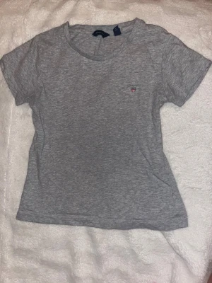 Grå t-shirt från GANT - Snygg grå t-shirt från GANT med liten broderad logga på bröstet. Klassisk rund hals och korta ärmar. Perfekt basic-plagg i mjuk bomull som funkar till jeans eller shorts. Enkel och stilren design som alltid är rätt.
