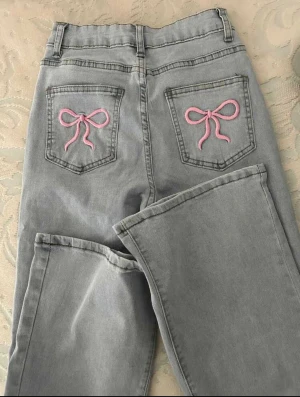 Blå jeans med rosa rosettbroderi - Snygga ljusblå jeans med bootcut och medelhög/låg midja. På bakfickorna finns broderade rosa rosetter som ger en gullig och unik touch. Jeansen är tillverkade i klassiskt denim och har en rak passform över låren som sedan blir vidare nedtill. Storlek L men passar även M! Sitter precis till hälen på mig som är ca 161cm! Helt nya men utan prislapp