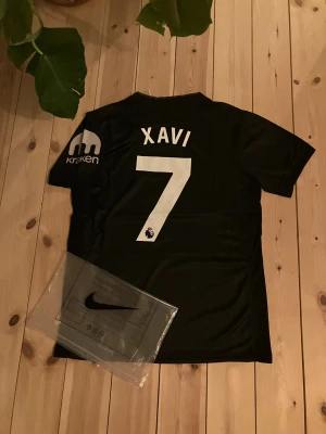 Tottenham Hotspur Nike fotbollströja svart - Säljer en svart Tottenham Hotspur fotbollströja från Nike med Dri-Fit material. Tröjan har vita tryck med AIA på bröstet, Spurs-logga och sponsor på ärmen. På ryggen står Xavi och nummer 7 i vitt. Perfekt för dig som älskar fotboll och Spurs.