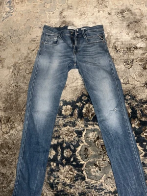 Replay Anbass blå jeans slim fit - Snygga Replay Anbass jeans i en klassisk blå tvätt med diskreta slitningar. Modellen har fem fickor, dragkedja och knappstängning. Slim fit med smalare ben och normal midja. Materialet är mjukt jeans-tyg i bomull med lite stretch för extra komfort. För fler frågor eller funderingar är det bara att höra av sig