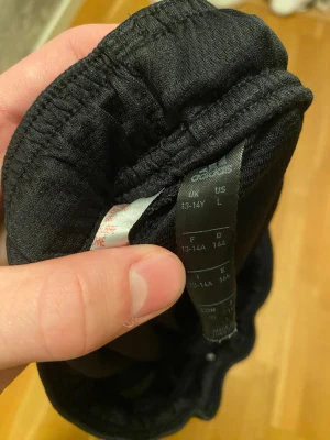 Svarta Adidas shorts med vita ränder - Svarta sportiga Adidas shorts i storlek L för tonåringar, med elastisk midja och vita detaljer. Ena benet har tre vita ränder och ett Adidas-märke. Materialet är lätt och syntetiskt, perfekt för träning eller fotboll. Snygg och enkel design med fickor på sidorna.