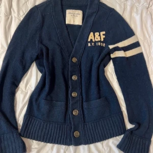 Marinblå kofta Abercrombie & Fitch 40% wool - Snygg marinblå stickad kofta från Abercrombie & Fitch med beige A&F-tryck och två vita ränder på ena ärmen. 2000s och tror om du gillar ”college” vibe är detta bra för dig! skriv för frågor, pris kan diskuteras 💕