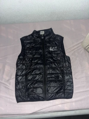 Svart dunväst från EA7 Emporio Armani - Snygg svart dunväst från EA7 Emporio Armani med quiltad design och hög krage. Västen har en tydlig logga på bröstet och dragkedja framtill. Perfekt att slänga över en hoodie för en cool streetstyle-look. Materialet är glansigt och känns lätt men ändå värmande. En jätte liten håll vid fickan typ fast den syns knappt tog mig ett tag tills jag hitta den (priset kan diskuteras) storlek S, en ny sån här väst kan ligga på värde mellan 1300-1800kr
