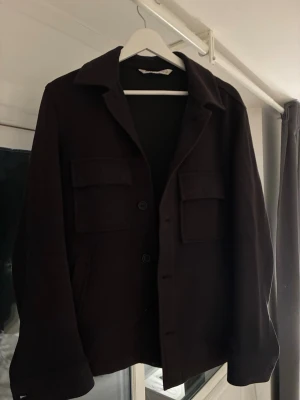 Svart overshirt - Stilren svart overshirt med klassisk krage och stora bröstfickor med lock. Jackan har knäppning med knappar framtill och raka ärmar. Perfekt för lager-på-lager och enkel att matcha med olika outfits.