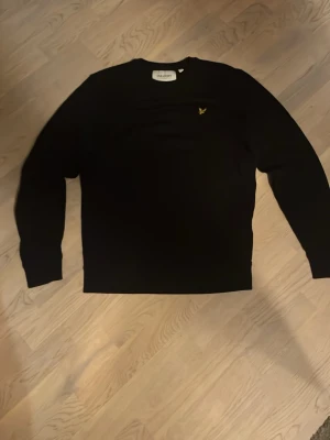 Svart tröja från Lyle & Scott - Svart långärmad tröja från Lyle & Scott med klassisk rund hals och gul broderad logga på bröstet. Tröjan har en clean och stilren look, perfekt för dig som gillar enkel design. Materialet känns mjukt och bekvämt mot huden.