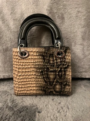 Beige handväska med ormmönster - Snygg liten handväska i beige med svart ormmönster och blanka svarta handtag. Väskan har en boxig form och detaljer i silverfärgad metall. Materialet är syntet med reptilstruktur som ger en cool look. Perfekt storlek för mobil och småsaker.