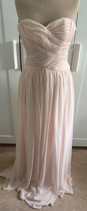 Ljusrosa långklänning från JS Boutique - Säljer en elegant ljusrosa långklänning från JS Boutique. Klänningen är axelbandslös med vackert draperad överdel och mjukt fallande kjol. Materialet är lätt och har en lyxig känsla, perfekt för speciella tillfällen. Passformen är normal och klänningen har en romantisk look.